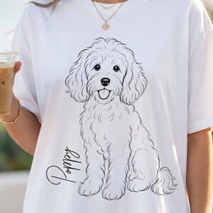 Può includere: Una t-shirt bianca a maniche corte con un'illustrazione a linee nere di un cane dal pelo folto seduto. Il nome Poppy è scritto in corsivo a sinistra del cane. Il disegno è centrato sulla parte anteriore del capo di abbigliamento.