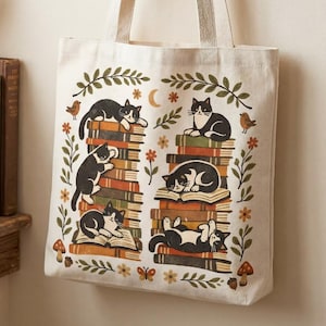 May include: Cream-colored tote bag featuring a whimsical design of black and white cats lounging on stacks of books. The illustration includes floral and celestial elements, creating a cozy, book-themed aesthetic. The bag has sturdy handles.