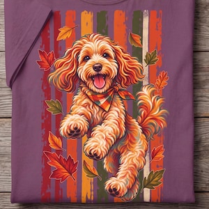 Puede incluir: Una camiseta color baya con un gráfico de un perro de pelaje rizado color marrón dorado que lleva un pañuelo a cuadros. El perro está rodeado de hojas de otoño que caen sobre un fondo de rayas verticales en tonos rojos, naranjas y verdes.