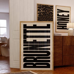 May include: A room interior featuring framed art prints. The largest print displays the word "BRUNETTE" in bold black letters against a white background. Other prints include a leopard print and the word "HONEY". The frames are light wood.