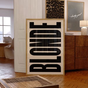 May include: A framed art print with the word "BLONDE" in large, bold, black letters. The print is in a light wood frame and is displayed in a room with other framed art pieces and wooden furniture. The room has a modern, minimalist aesthetic.