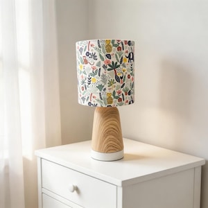 Peut inclure: Une lampe de table décorative avec un abat-jour cylindrique orné d'un imprimé floral et animalier coloré. La lampe a un socle en bois avec un fond blanc et repose sur une table de chevet blanche. L'arrière-plan comprend une fenêtre avec des rideaux blancs transparents.