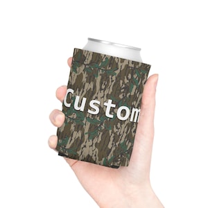 May include: A cylindrical foam can cooler featuring a brown, green, and black tree bark camouflage pattern. The word Custom is printed in large, bold, white sans-serif capital letters across the center of the exterior surface of the cooler.