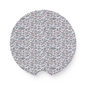May include: A round car coaster with a camouflage pattern in shades of gray, brown, and white. The coaster has a small notch on one side for easy removal from a cup holder. The design is repeated across the surface.