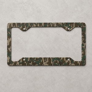 May include: License plate frame with a camouflage pattern in shades of brown, green, and tan. The frame has a rectangular shape with rounded corners and four mounting holes. The camouflage design is a repeating pattern.