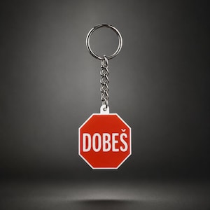 May include: A red and white octagonal keychain with the word "DOBEŠ" in white letters. The keychain has a silver chain and ring for attaching to keys or other items. The background is a gradient of dark grey.