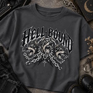 Könnte beinhalten: Ein anthrazitfarbenes T-Shirt mit einem Grafikdruck von drei knurrenden Dobermann-Köpfen, die mit Stachelhalsbändern versehen und durch silberne Ketten verbunden sind. Der Text HELL BOUND steht in einer großen, zackigen Gothic-Schrift darüber.