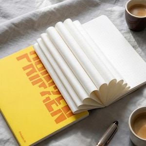 Puede incluir: Cuaderno amarillo abierto con la palabra "PRINZEN" en letras naranja. Las páginas blancas tienen pequeños puntos grises. Un bolígrafo y dos tazas de café se ven en el fondo.