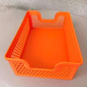 May include: Bright orange plastic desk organizer with a rectangular shape. The sides have a perforated design, and the front has a curved cutout for easy access. The organizer is designed to hold papers or small office supplies.
