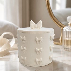 May include: A cream-colored, round container with a lid, adorned with bow-shaped embellishments. The lid features a single bow, and the sides are decorated with multiple bows. The container is likely used for storage or decoration.