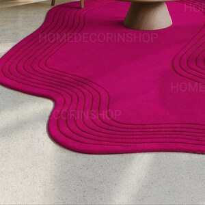 Op de afbeelding: Een levendig magenta tapijt met een unieke, vloeiende vorm. Het tapijt heeft een reeks gebogen, verhoogde lijnen die een textuureffect cre&euml;ren. Het tapijt ligt op een lichtgekleurde betonnen vloer, met een houten tafel op de achtergrond. De tekst "HOMEDECORINSHOP" is zichtbaar.
