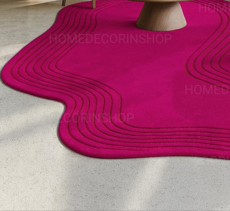 Op de afbeelding: Een levendig magenta tapijt met een unieke, vloeiende vorm. Het tapijt heeft een reeks gebogen, verhoogde lijnen die een textuureffect cre&euml;ren. Het tapijt ligt op een lichtgekleurde betonnen vloer, met een houten tafel op de achtergrond. De tekst "HOMEDECORINSHOP" is zichtbaar.