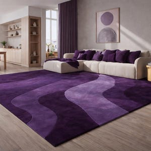 Può includere: Un grande tappeto rettangolare viola con un design ondulato in varie tonalità di viola. Il tappeto è posizionato su un pavimento in legno in un soggiorno. È visibile un divano angolare bianco con cuscini viola.
