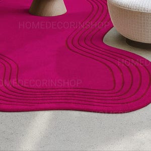 Op de afbeelding: Een levendig magenta tapijt met een uniek, vloeiend ontwerp. Het tapijt heeft een reeks gebogen, verhoogde lijnen in een donkerdere tint magenta, waardoor een textuureffect ontstaat. De vorm van het tapijt is organisch en onregelmatig, wat een moderne touch aan de kamer toevoegt. De tekst "HOMEDECORINSHOP" is zichtbaar.