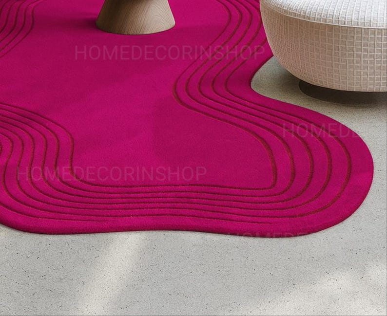 Op de afbeelding: Een levendig magenta tapijt met een uniek, vloeiend ontwerp. Het tapijt heeft een reeks gebogen, verhoogde lijnen in een donkerdere tint magenta, waardoor een textuureffect ontstaat. De vorm van het tapijt is organisch en onregelmatig, wat een moderne touch aan de kamer toevoegt. De tekst "HOMEDECORINSHOP" is zichtbaar.
