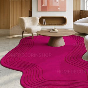 Op de afbeelding: Een levendig magenta tapijt met een uniek, vloeiend ontwerp. Het tapijt heeft een reeks gebogen lijnen, waardoor een golfachtig patroon ontstaat. Een ronde houten salontafel en een cr&egrave;mekleurige fauteuil staan op de achtergrond. De tekst "HOMEDECORINSHOP" is zichtbaar.
