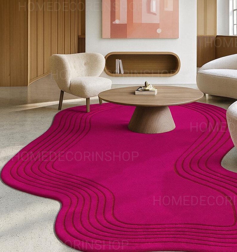 Op de afbeelding: Een levendig magenta tapijt met een uniek, vloeiend ontwerp. Het tapijt heeft een reeks gebogen lijnen, waardoor een golfachtig patroon ontstaat. Een ronde houten salontafel en een cr&egrave;mekleurige fauteuil staan op de achtergrond. De tekst "HOMEDECORINSHOP" is zichtbaar.