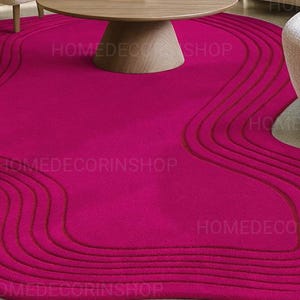 Op de afbeelding: Een levendig magenta vloerkleed met een uniek, vloeiend ontwerp. Het vloerkleed heeft een reeks gebogen, verhoogde lijnen in een donkerdere tint magenta, waardoor een textuurachtig, golfachtig patroon ontstaat. Het vloerkleed ligt onder een houten salontafel en in de buurt van een witte fauteuil.