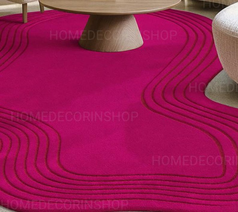 Op de afbeelding: Een levendig magenta vloerkleed met een uniek, vloeiend ontwerp. Het vloerkleed heeft een reeks gebogen, verhoogde lijnen in een donkerdere tint magenta, waardoor een textuurachtig, golfachtig patroon ontstaat. Het vloerkleed ligt onder een houten salontafel en in de buurt van een witte fauteuil.