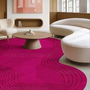 Op de afbeelding: Een levendig magenta vloerkleed met een uniek, vloeiend ontwerp. Het vloerkleed heeft een reeks gebogen lijnen in een donkerdere tint magenta, waardoor een gelaagd effect ontstaat. Het vloerkleed staat in een moderne woonkamer met een ronde houten salontafel en een gebogen bank.