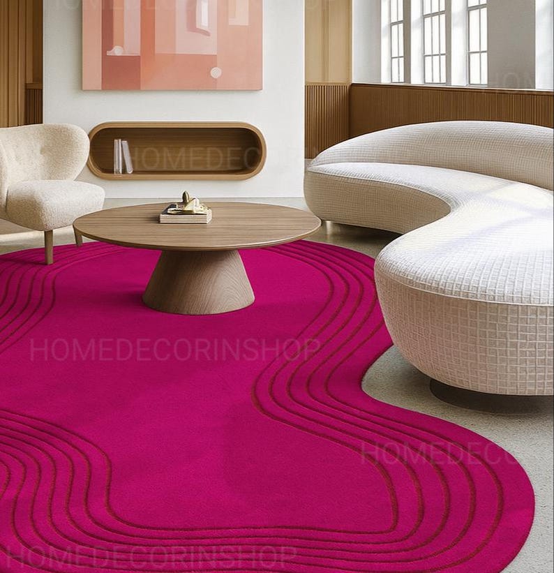 Op de afbeelding: Een levendig magenta vloerkleed met een uniek, vloeiend ontwerp. Het vloerkleed heeft een reeks gebogen lijnen in een donkerdere tint magenta, waardoor een gelaagd effect ontstaat. Het vloerkleed staat in een moderne woonkamer met een ronde houten salontafel en een gebogen bank.