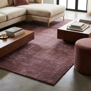 May include: A rectangular area rug in a deep mauve color with a soft, textured pile. The rug is positioned on a concrete floor beneath a beige sectional sofa, two wooden coffee tables with stacked books, and a matching mauve velvet ottoman.
