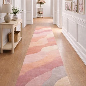 Pode incluir: Um tapete longo e retangular com um design ondulado em tons de rosa, pêssego e cinza. O tapete está colocado num corredor com paredes claras e chão de madeira. As dimensões do tapete não são visíveis.
