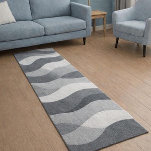 Può includere: Un tappeto rettangolare con un motivo a onde in tonalità di grigio e bianco. Il tappeto è posizionato su un pavimento in legno, di fronte a un divano e una poltrona azzurri, creando un'estetica neutra e rilassante.