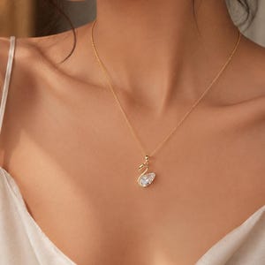 May include: A gold-toned necklace featuring a delicate chain and a swan-shaped pendant. The swan pendant is outlined in gold metal and filled with a clear, pear-shaped faceted gemstone that catches the light, creating a subtle sparkle.