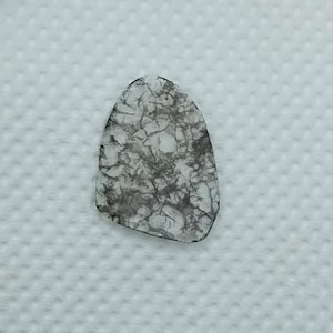 May include: A translucent, teardrop-shaped stone with a complex, web-like pattern of grey and black veins. The stone is set against a white, textured background, highlighting its unique internal structure and natural beauty.