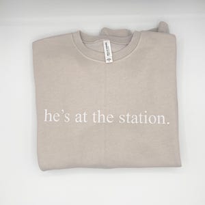 May include: A beige crew neck sweatshirt with the text "he's at the station" printed in white. The sweatshirt is folded. The fabric appears to be soft and comfortable. A tag is attached to the collar.