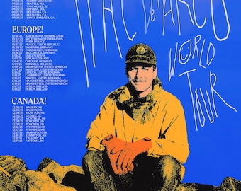 Op de afbeelding: Poster voor Mac DeMarco's World Tour, met een foto van de artiest in een gele jas en pet. Tourdata staan vermeld voor de Verenigde Staten, Europa en Canada. De achtergrond is blauw.