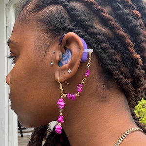 May include: A side profile view of a person wearing a hearing aid with a purple and gold chain earring. The hearing aid is blue and white. The earring features pink gemstone beads. The person has dark brown dreadlocks and a gold chain necklace.