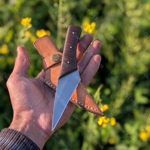 以下が含まれることがあります： 濃い木製の柄と銀色の刃を持つ手作りのナイフ。茶色の革製の鞘が付属しています。ナイフはユニークな角度のあるデザインです。鞘には装飾的な金属製の留め金が付いています。背景は緑の葉のぼやけた景色です。