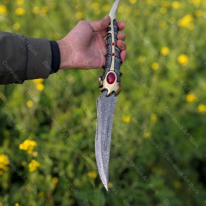 May include: An ornate dagger with a silver blade and a decorative handle featuring gold, red, and silver accents. The handle has a detailed design with a large red gem. The blade has a textured pattern.
