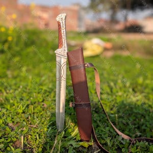 以下が含まれることがあります： 円形のラインパターンが刻まれた刃と、複雑な銀色の金属象嵌が施された茶色の木製ハンドルを持つ銀色の金属製短剣。この短剣には、一致するレザーストラップが付いた茶色のレザーシースが付属しています。