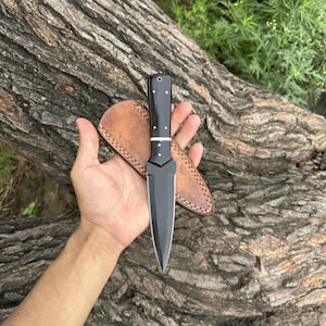 May include: A black dagger with a polished blade and a black handle with white accents. The dagger is paired with a brown leather sheath with decorative stitching. The knife is approximately 10 inches long.
