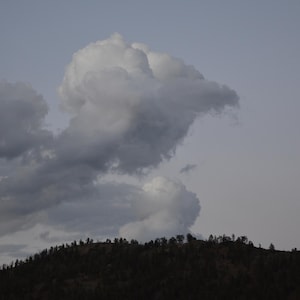 May include: A dramatic landscape featuring a cloudy sky and a dark, tree-covered mountain range. The clouds are a mix of gray and white, with a large, puffy formation dominating the center. The overall scene evokes a sense of nature and the outdoors.