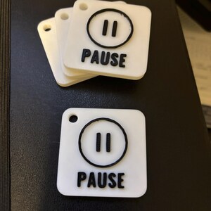 May include: White square tags with a black pause symbol and the word "PAUSE" printed in black. The tags have a small hole at the top for attaching to a keychain or lanyard. Several tags are stacked together.