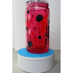 Op de afbeelding: Een heldere, rood getinte glazen beker met een doorzichtig deksel. De beker is versierd met zwarte rozen, harten en een skeletontwerp. De beker staat op een wit en blauw oppervlak.