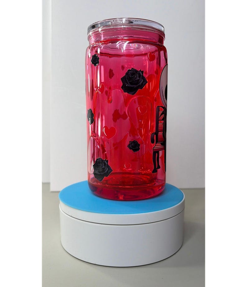 Op de afbeelding: Een heldere, rood getinte glazen beker met een doorzichtig deksel. De beker is versierd met zwarte rozen, harten en een skeletontwerp. De beker staat op een wit en blauw oppervlak.