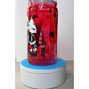 Op de afbeelding: Een heldere glazen pot gevuld met rode vloeistof, met een cartoon illustratie van een meisje met rood haar en een zwarte jurk. De pot heeft een deksel en zwarte rozen. De pot staat op een blauw en witte draaiende standaard.