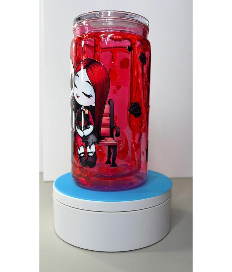 Op de afbeelding: Een heldere glazen pot gevuld met rode vloeistof, met een cartoon illustratie van een meisje met rood haar en een zwarte jurk. De pot heeft een deksel en zwarte rozen. De pot staat op een blauw en witte draaiende standaard.