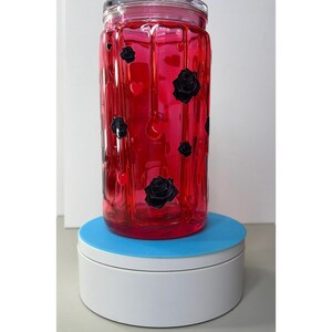 Op de afbeelding: Een heldere glazen pot gevuld met een levendige rode vloeistof. De pot is versierd met zwarte rozen en hartvormige stickers. De pot staat op een witte en blauwe roterende standaard.