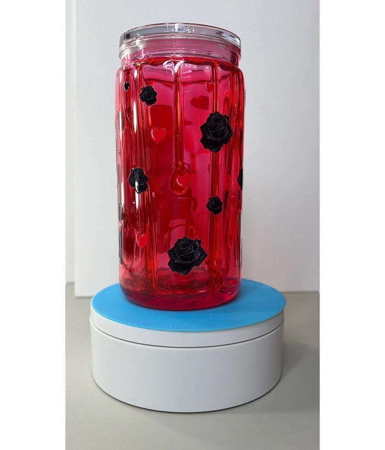 Op de afbeelding: Een heldere glazen pot gevuld met een levendige rode vloeistof. De pot is versierd met zwarte rozen en hartvormige stickers. De pot staat op een witte en blauwe roterende standaard.
