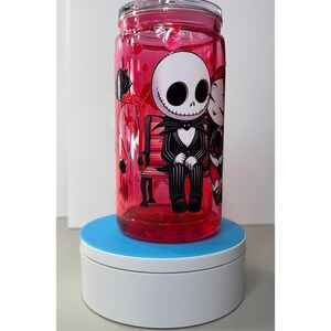 Op de afbeelding: Een heldere, rood getinte glazen beker met een deksel, met een cartoonillustratie van Jack Skellington en Sally uit The Nightmare Before Christmas. De personages zitten op een bank. De beker is gevuld met een rode vloeistof en hartvormige confetti.