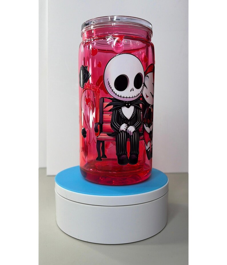 Op de afbeelding: Een heldere, rood getinte glazen beker met een deksel, met een cartoonillustratie van Jack Skellington en Sally uit The Nightmare Before Christmas. De personages zitten op een bank. De beker is gevuld met een rode vloeistof en hartvormige confetti.