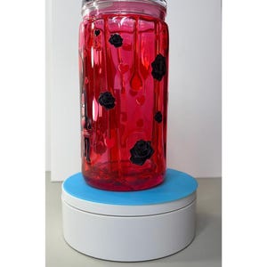 Op de afbeelding: Een heldere, rode glazen pot met een deksel, versierd met zwarte rozen en hartvormige accenten. De pot is cilindrisch en staat op een wit en blauw draaiplateau. De pot heeft een gestructureerd oppervlak.