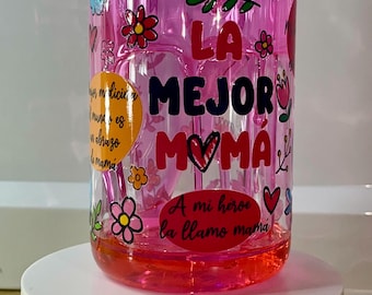 May include: A clear glass jar with a pink liquid inside, decorated with colorful floral and butterfly designs. The jar has text in Spanish that reads "LA MEJOR MAMA" and "A mi hero la llamo mama". The jar sits on a white pedestal.