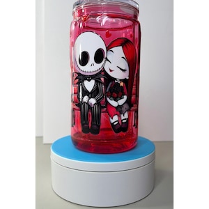 Op de afbeelding: Een heldere, cilindrische beker met een rode vloeistof erin en een schroefdeksel. De beker heeft een cartoonillustratie van Jack Skellington en Sally uit The Nightmare Before Christmas. De personages zitten op een bank, omringd door harten.